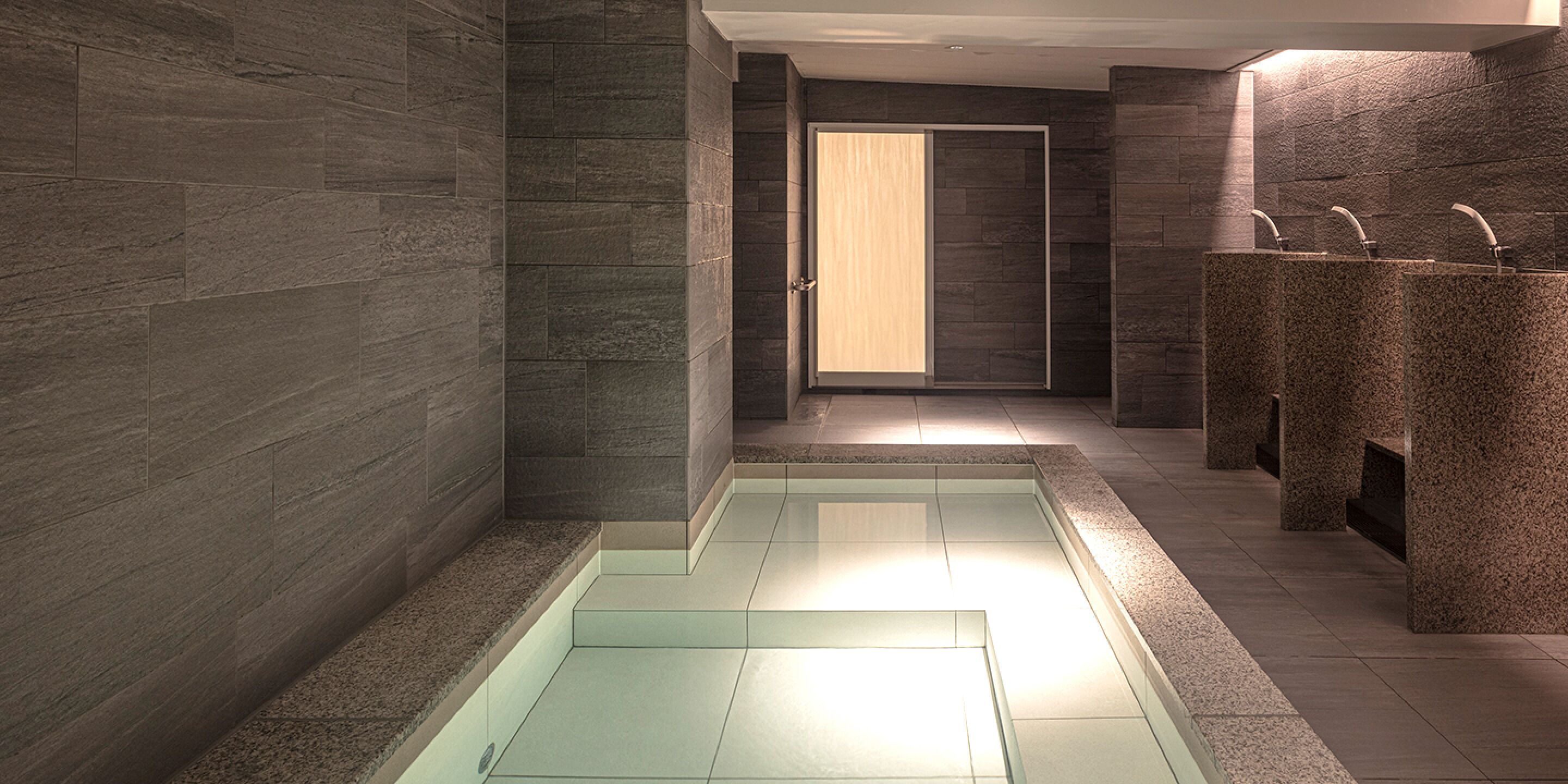 A modern bathroom with a glass-enclosed pool, dark stone walls, and three granite sinks.