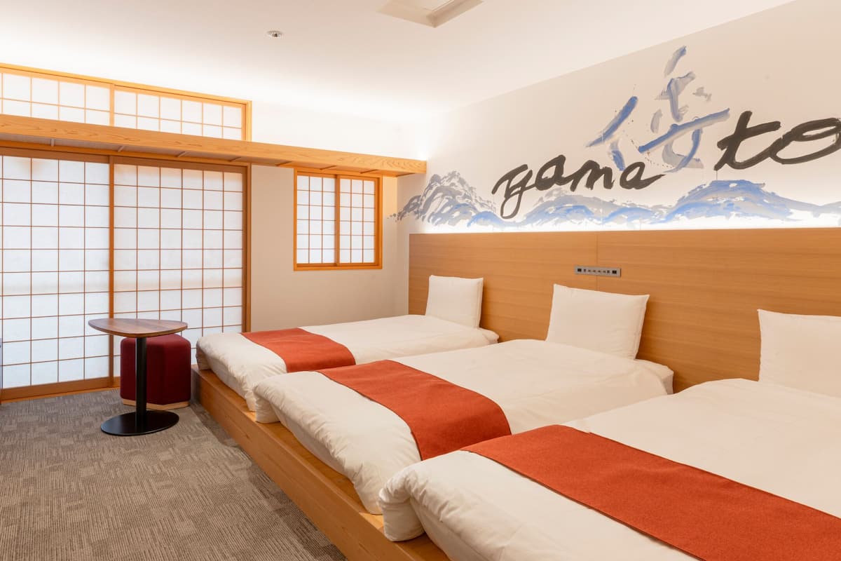 A hotel room with two beds, a small table, a red ottoman, and a view of a mountain through a shoji screen.