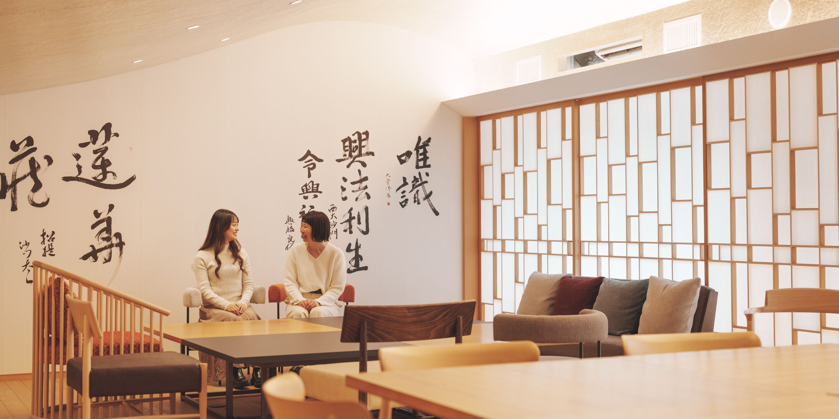 A room with a wooden table and chairs, a white wall with black calligraphy, and a traditional wooden screen.