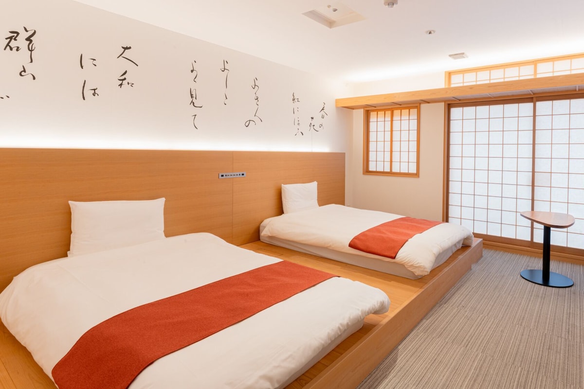 Habitación de hotel con dos camas, una mesita y vistas a una ventana tradicional japonesa.