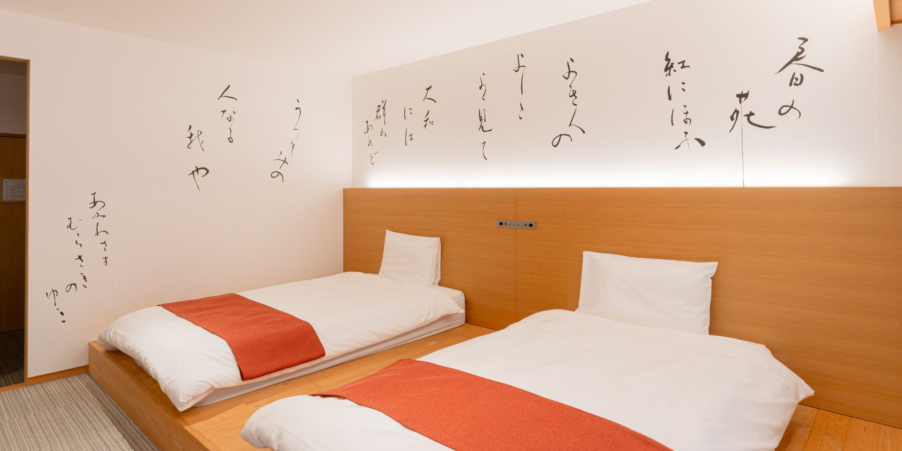 A hotel room with two beds, each with white bedding and orange throw pillows. The room features a wooden headboard and a wall with Japanese calligraphy.