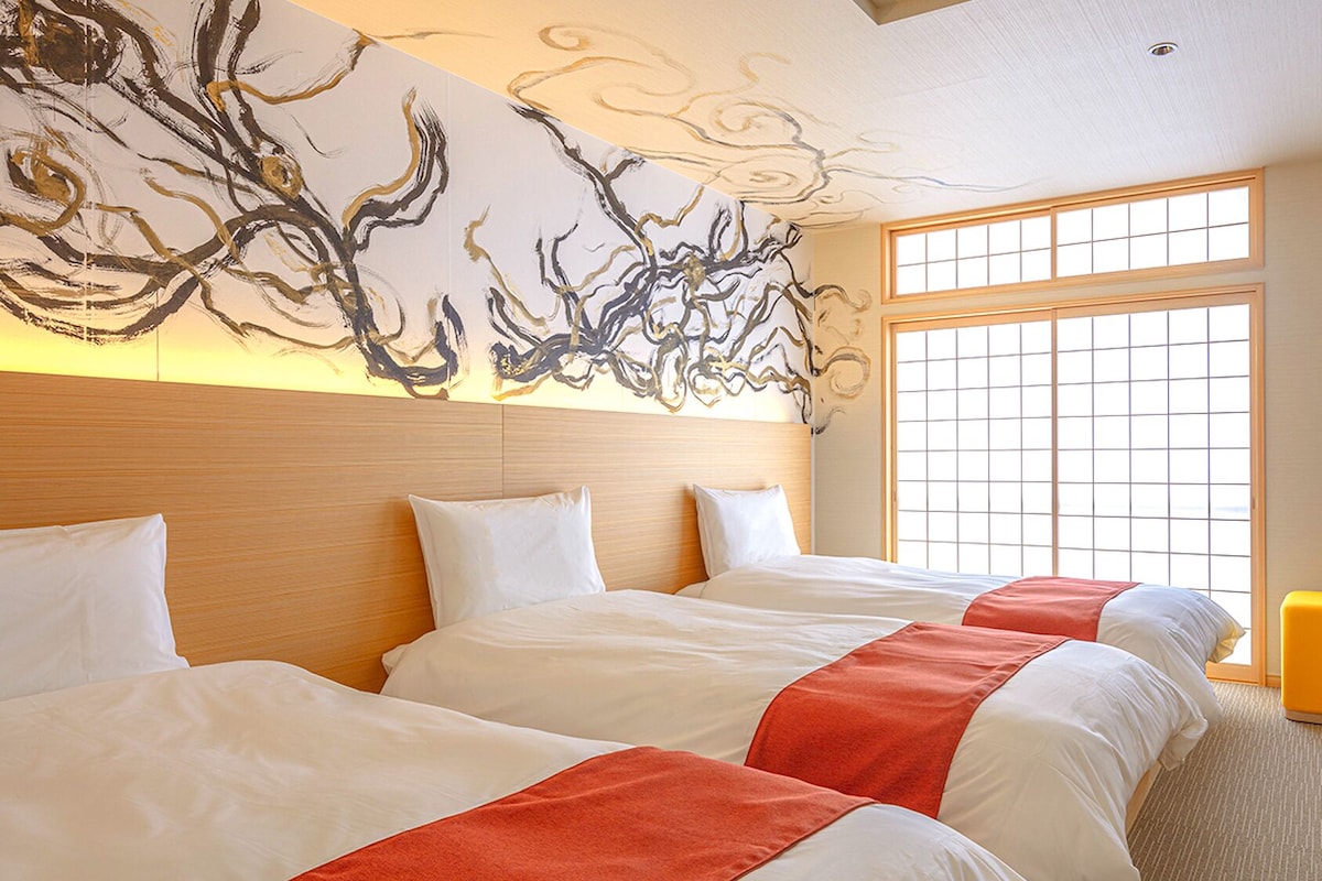 Habitación de hotel con dos camas, un cuadro abstracto grande en la pared, una ventana con diseño de cuadrícula y una pequeña mesa redonda con un taburete amarillo.