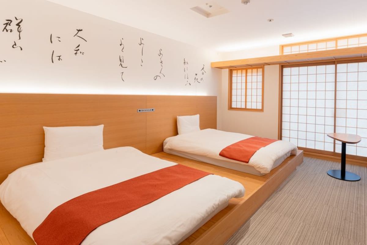 Habitación de hotel con dos camas, una mesita y vistas a través de un celosía.