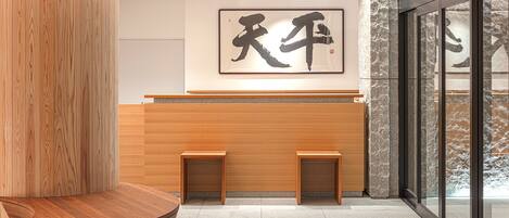 A reception area with a wooden reception desk, two small side tables, a large calligraphy artwork on the wall, and a stone wall to the right.
