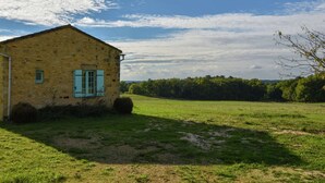 Un rústico edificio de piedra con una sola ventana y una puerta, rodeado de un campo de hierba y árboles al fondo.