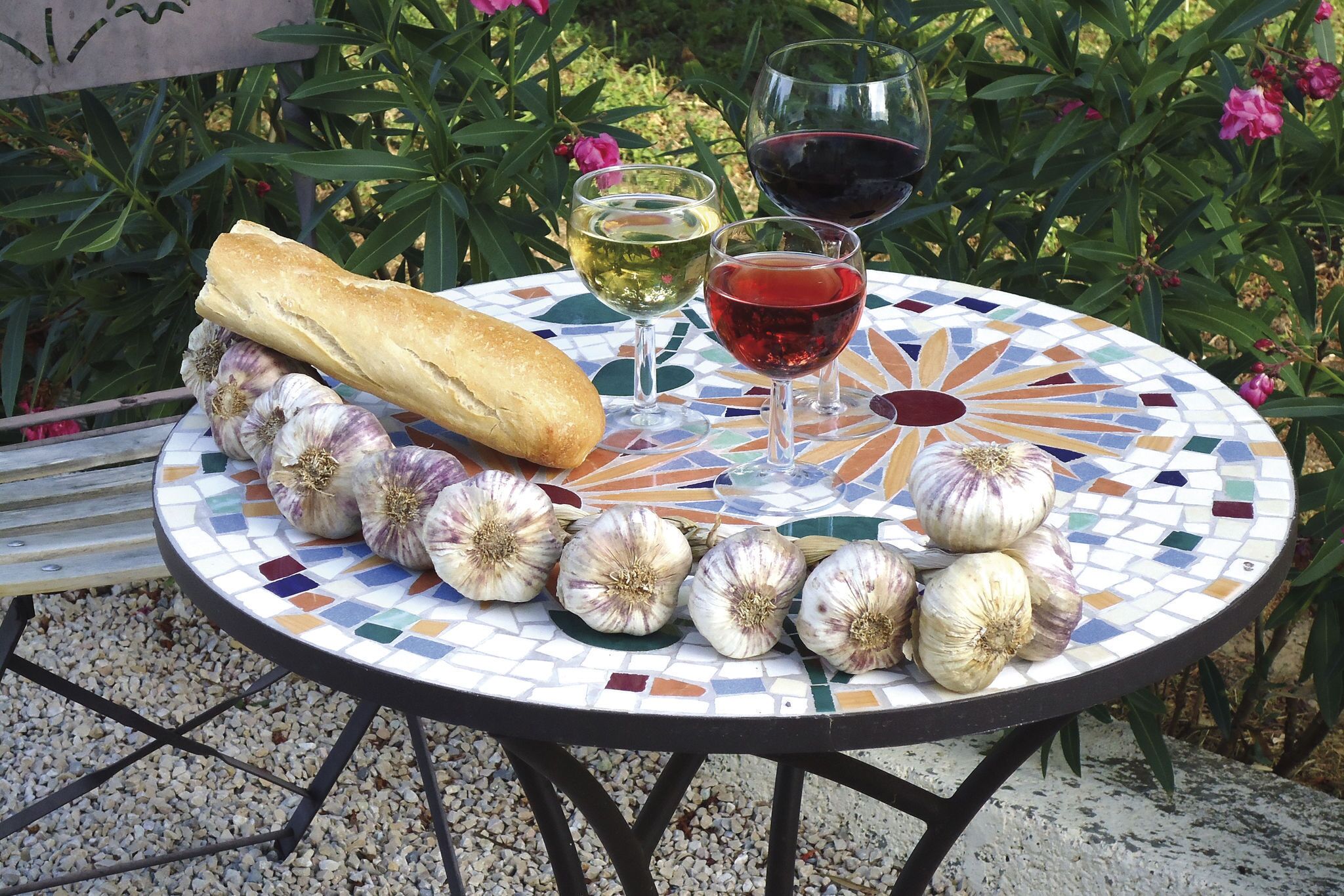 A round table with a mosaic pattern, a baguette, garlic bulbs, and two wine glasses filled with white and red wine.