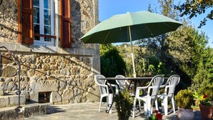 A stone building with a wooden door and window shutters, a stone wall, and a patio area with a table and chairs under a green umbrella. - Sénezergues Garden Hideaway (Sénezergues)