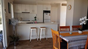 A modern kitchen with white cabinets, a microwave, and a refrigerator. There is a dining area with a table and chairs, and a potted plant on the floor. - Omeo Mountain Views. Peace and Tranquillity (Omeo)