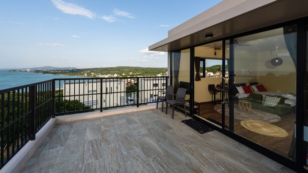 Ein Balkon mit Meerblick, eine Glastür zu einem Wohnbereich mit einer Couch und Sesseln sowie ein kleiner Teppich auf dem Boden.