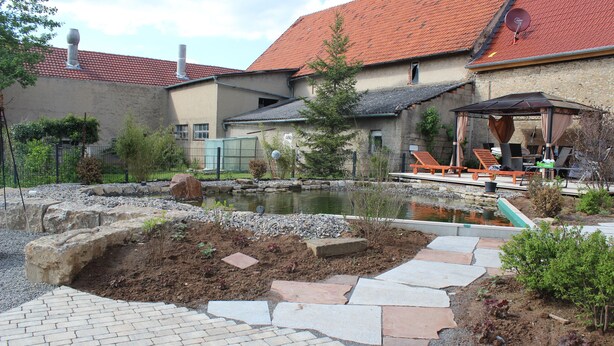 A house with a backyard containing a pond, a patio with lounge chairs, and a stone pathway.