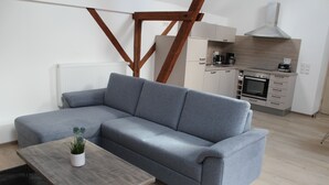 A modern living room with a grey sectional sofa, a wooden coffee table with a plant, and a kitchen in the background with a built-in oven and microwave.