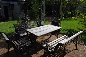 A patio with a wooden table and benches, surrounded by a garden with a fence and a pathway. - Luxury Heritage Cottage In The Heart Of Town (Mittagong)