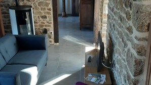 A living room with a stone wall, a sofa, a coffee table with magazines, and a TV on a stand. - Charming stone house in Villedieu with garden (Villedieu-les-Poêles)