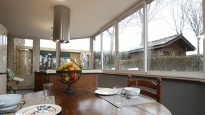 A dining area with a wooden table, a fruit bowl, and a view of a house through large windows. - Charming stone house in Villedieu with garden (Villedieu-les-Poêles)