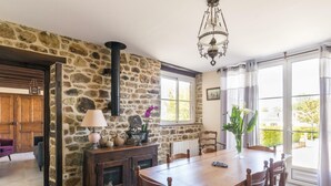 A rustic dining area with a stone wall, wooden furniture, and a large window with curtains. - Charming stone house in Villedieu with garden (Villedieu-les-Poêles)