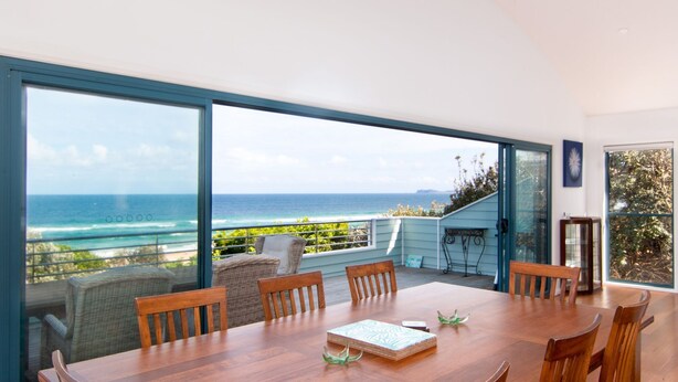 A dining area with a view of the ocean, a wooden table, and wicker chairs. - Huey's Lookout - beachfront spacious pet friendly home (Blueys Beach)