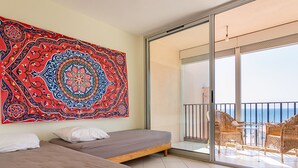 Une chambre d’hôtel avec deux lits, une grande tapisserie aux couleurs vives et un balcon meublé de mobilier en osier, donnant sur la mer.