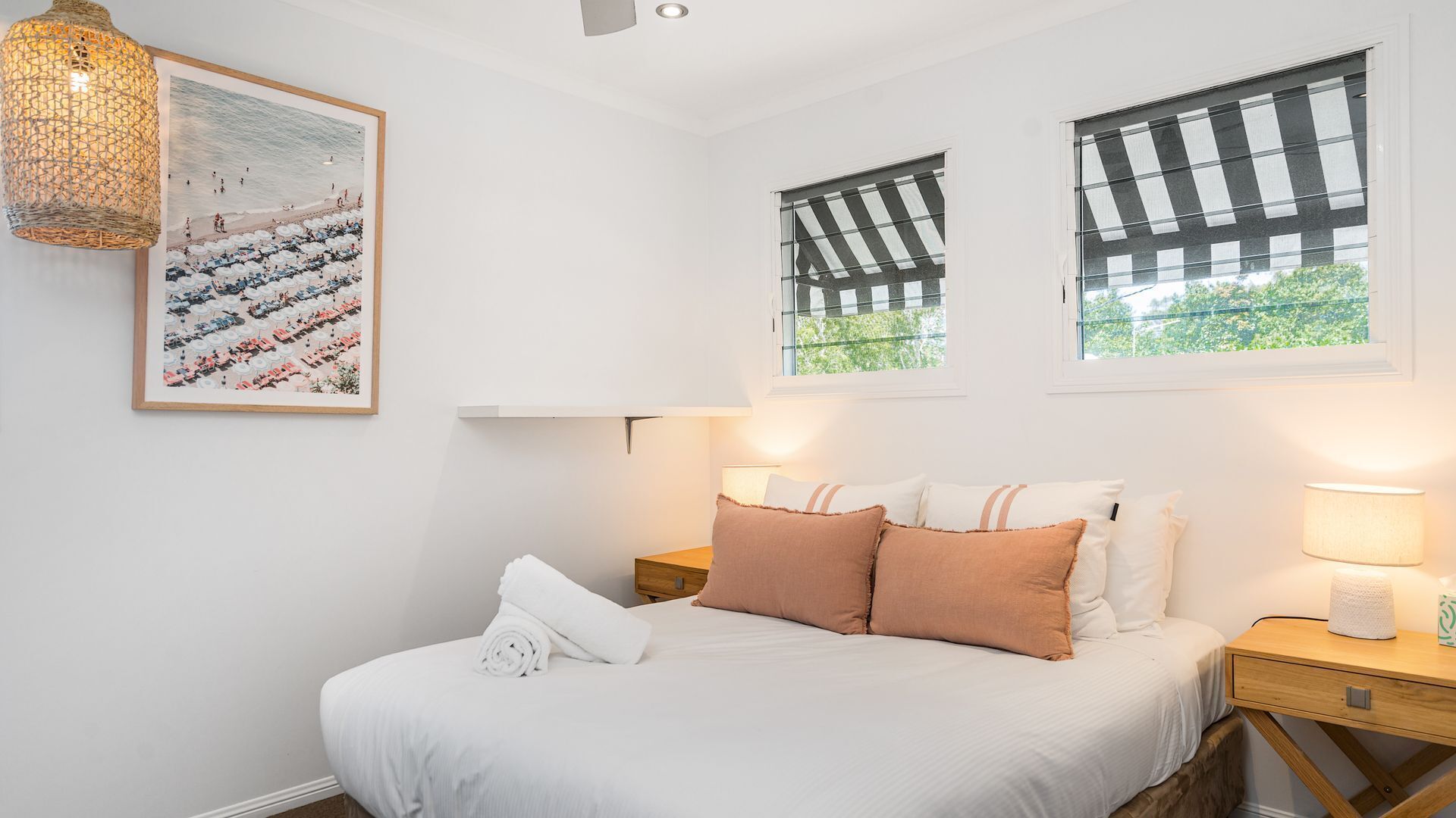 A bedroom with a bed, pillows, a nightstand, a lamp, a wall-mounted picture, and a window with blinds.