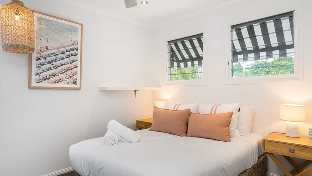 A bedroom with a bed, pillows, a nightstand, a lamp, a wall-mounted picture, and a window with blinds.