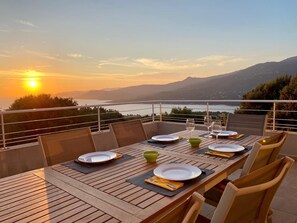 Une table à manger dressée pour quatre personnes, avec assiettes, bols et couverts, offrant une vue panoramique sur un lac et des montagnes au coucher du soleil.