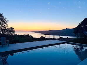 Une piscine offrant une vue sur une étendue d’eau et sur des montagnes lointaines, au coucher du soleil.