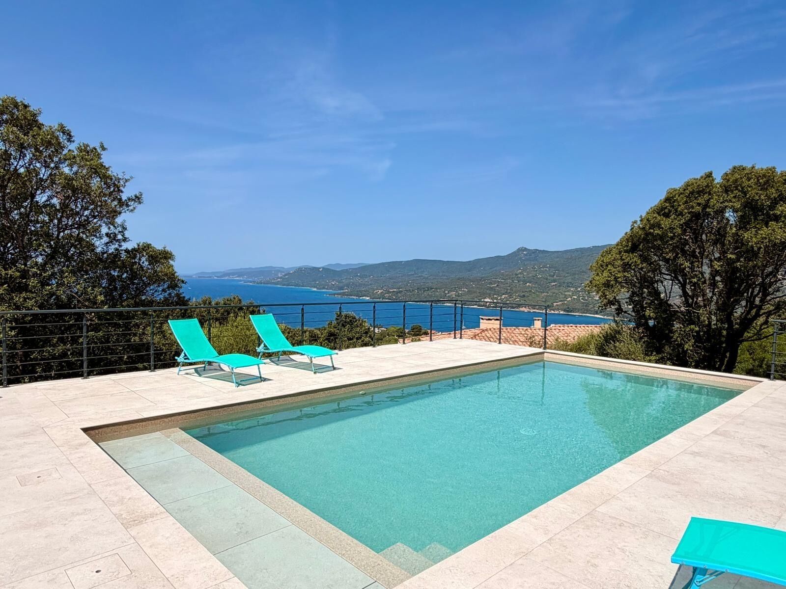 Et rektangulært pool med to blå liggestole på den ene side, omgivet af en flisbelagt terrasse, med udsigt til havet og bakker i baggrunden.