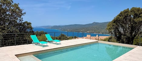 Una piscina rectangular con dos sillas reclinables azules a un lado, rodeada por un solárium con baldosas, y vistas al mar y colinas al fondo.