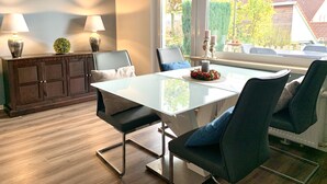 Ein moderner Essbereich mit einem Glastisch, dunklen Stühlen, einem Holzkabinett mit Lampen und Blick ins Freie.