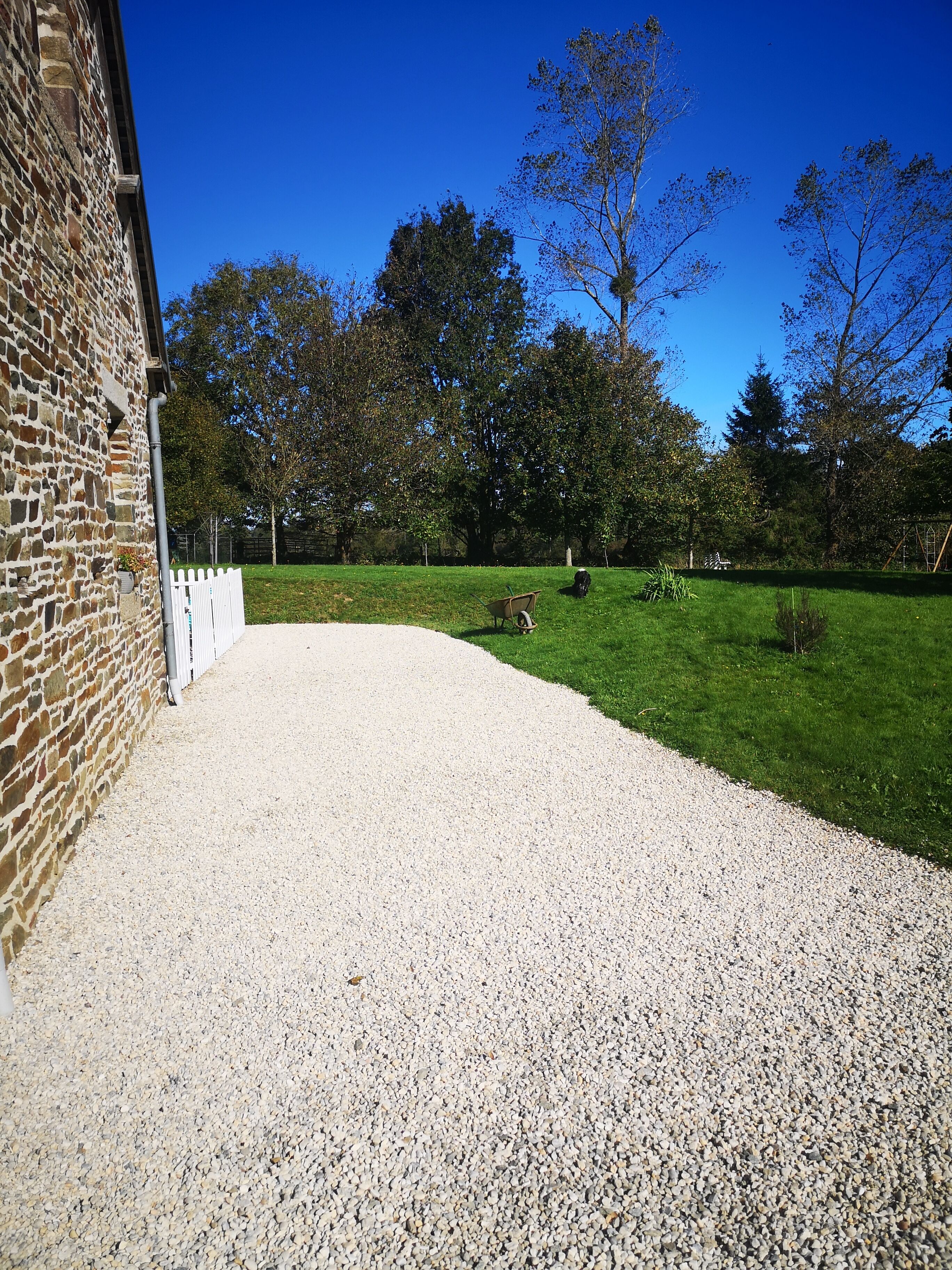 A gravel driveway leading to a white picket fence, a stone building, and a grassy area with trees.