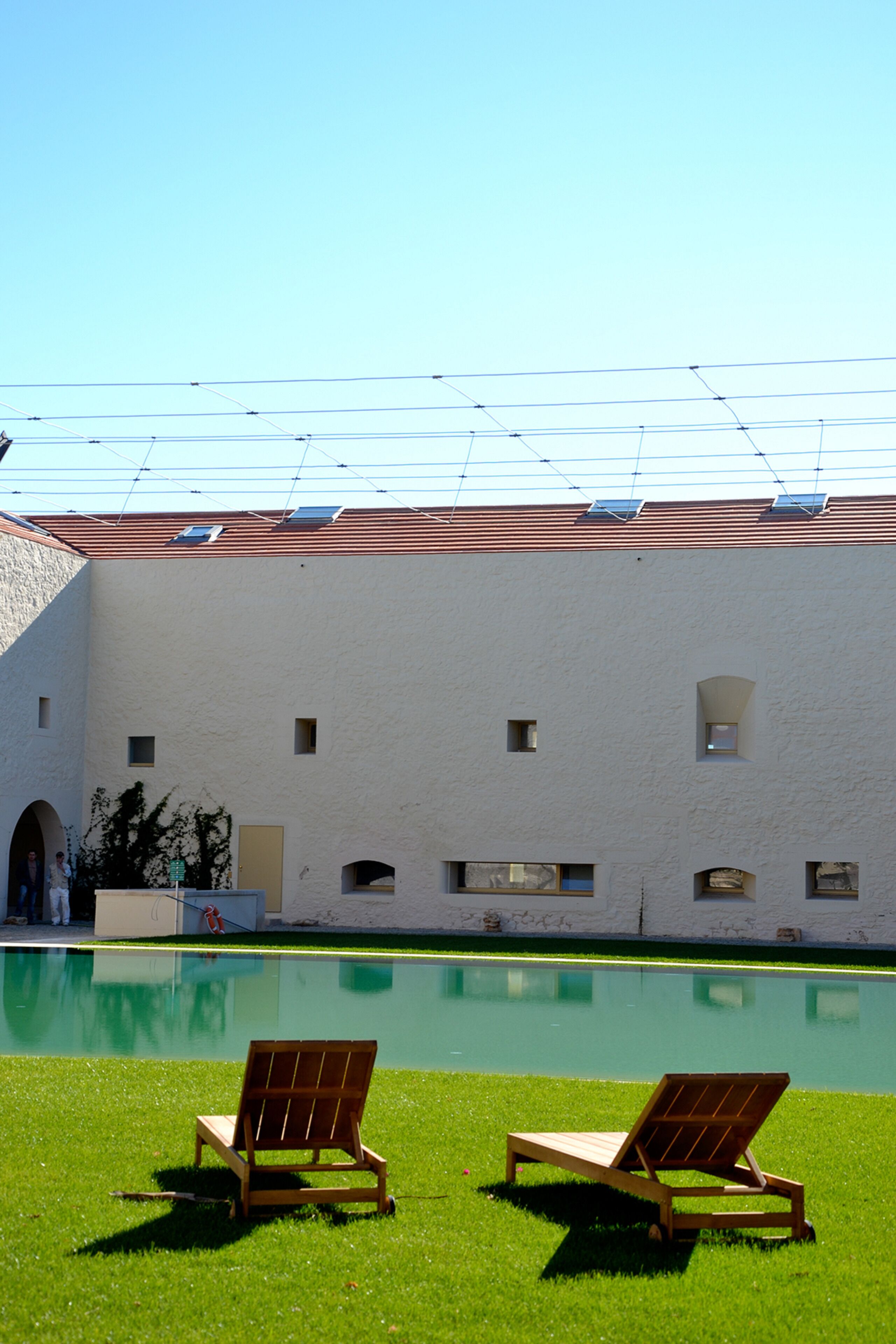 A poolside area with two wooden lounge chairs and a building with a tiled roof and small windows.