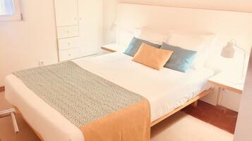 Una cama bien tendida con una colcha y almohadas estampadas, una mesita de noche con lámpara y un armario en una habitación luminosa.