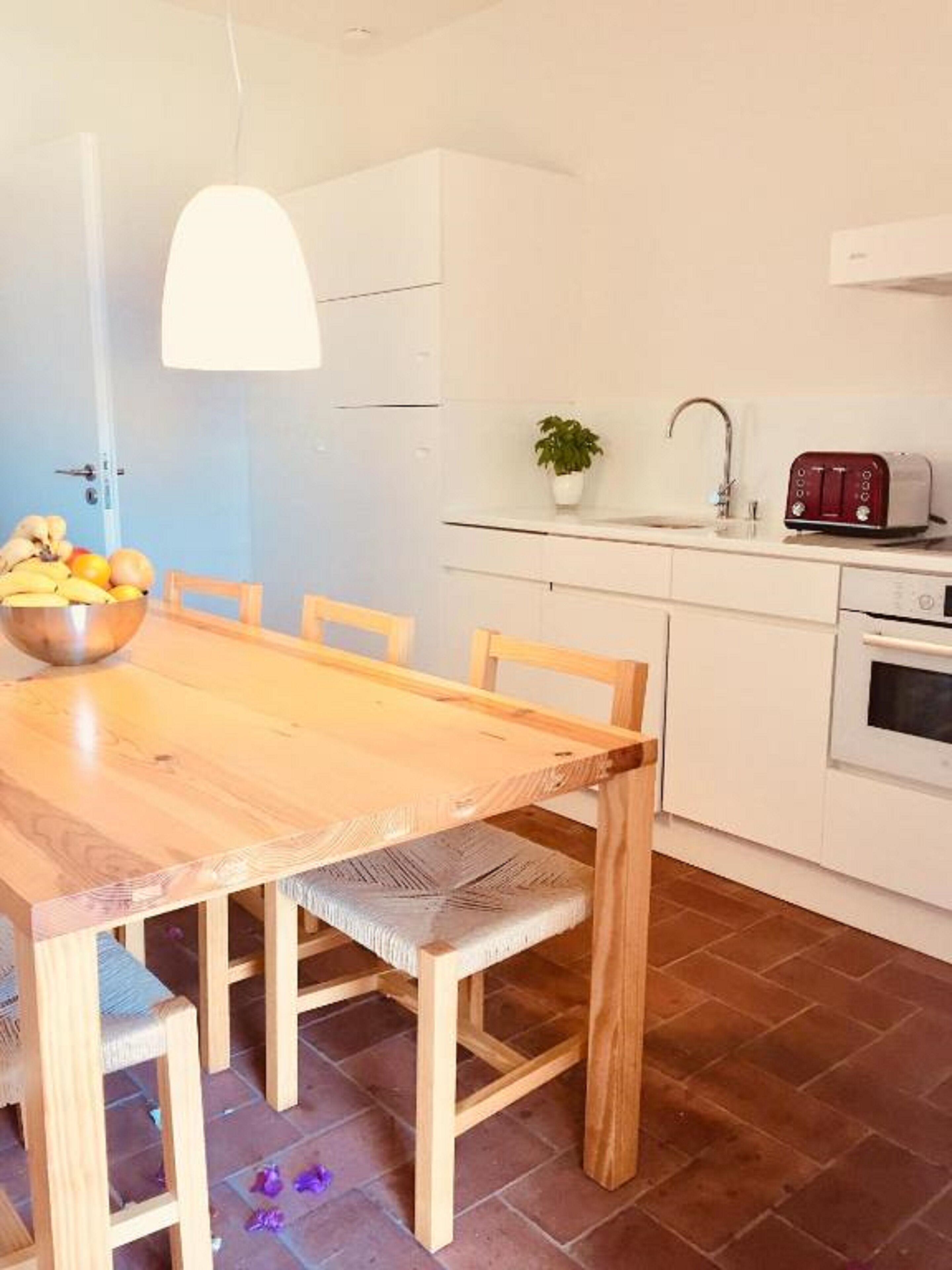 A wooden dining table with chairs, a bowl of fruit, a potted plant, a toaster, and a sink in a kitchen area.