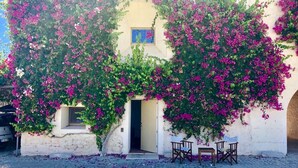 A building with a white facade and pink flowering vines. There is a doorway leading to an interior space with a small window and a table with chairs outside.
