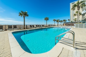 A swimming pool with a clear blue water, surrounded by lounge chairs and palm trees, with a building in the background.