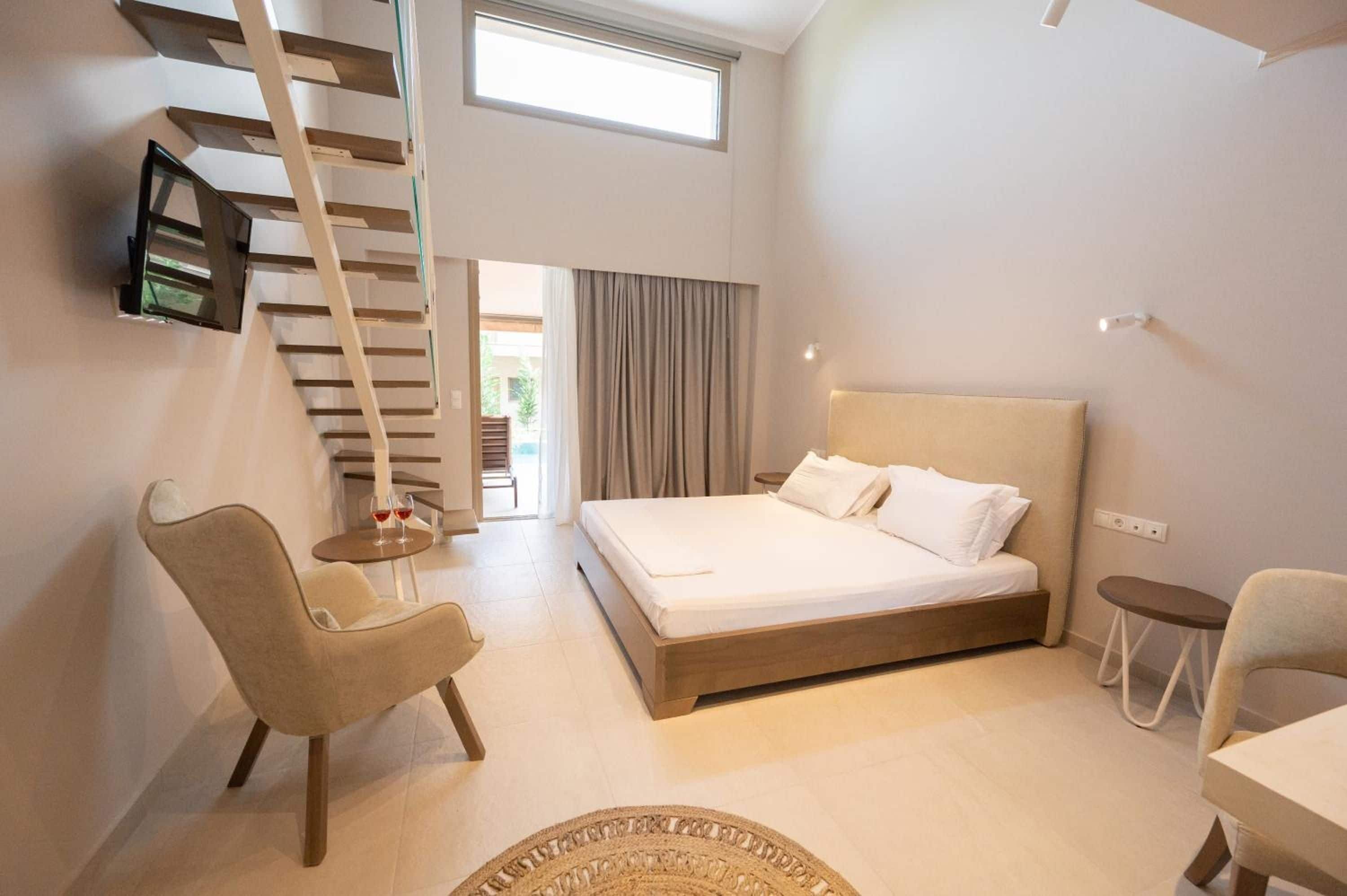 A modern bedroom with a large bed, a chair, a small table, a TV mounted on the wall, and a staircase.