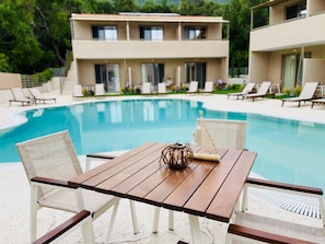 A poolside table with two chairs, a decorative item, and a building with multiple windows in the background. - Scarabeo Suites & Villas (Parga)