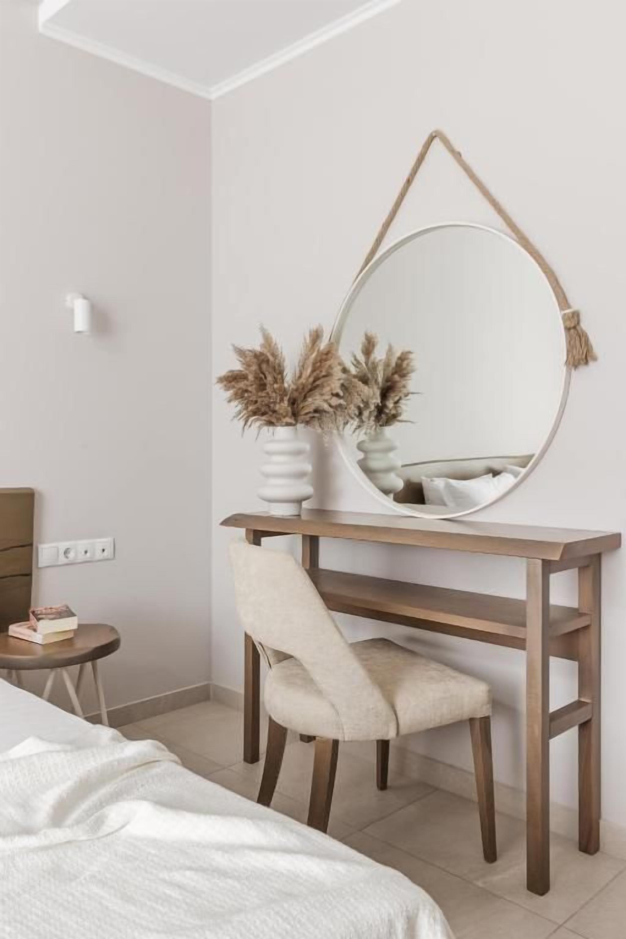 A bedroom with a bed, a desk with a chair, a round mirror, and a vase with dried plants.