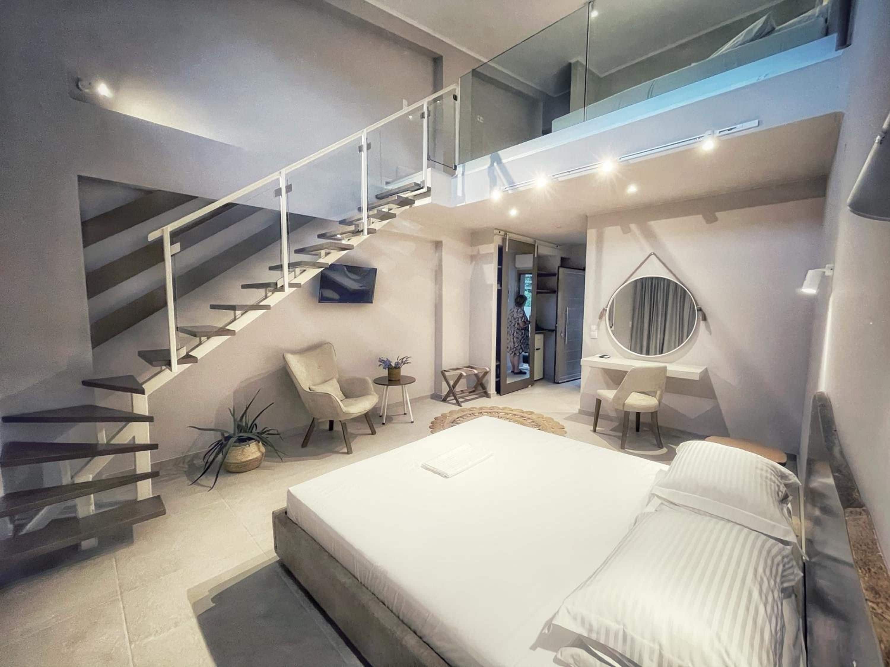 A modern bedroom with a large bed, a desk, a chair, a small table, a round mirror, and a staircase.