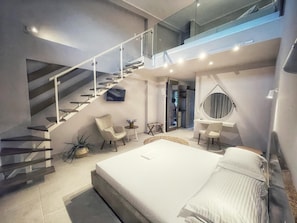 A modern bedroom with a large bed, a desk, a chair, a small table, a round mirror, and a staircase. - Scarabeo Suites & Villas (Parga)