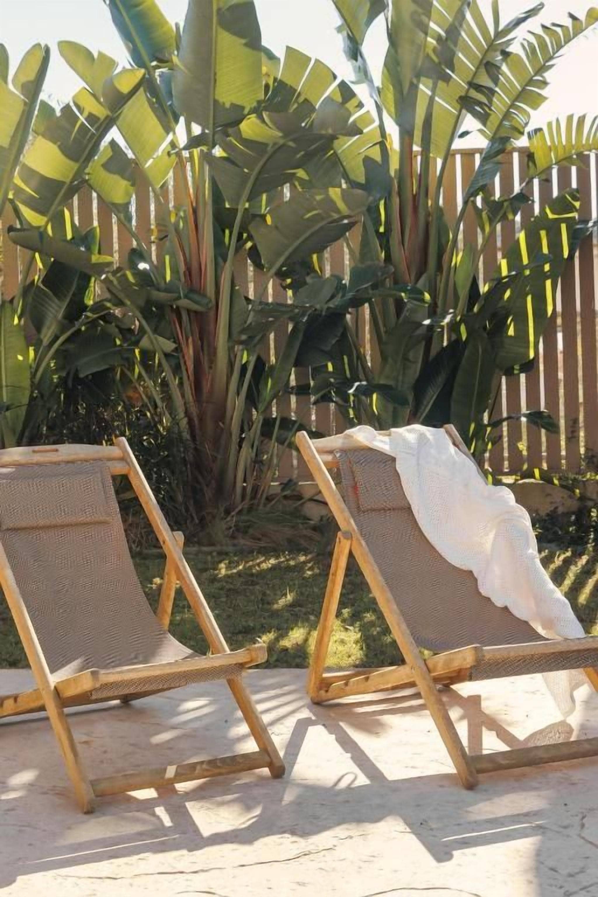 Two wooden lounge chairs with mesh backrests, positioned on a sandy surface with a wooden fence and banana plants in the background.