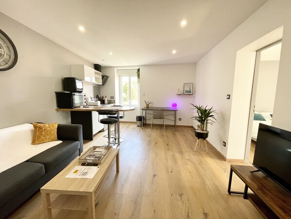 A modern living room with a sofa, coffee table, and TV stand. A kitchen area with a dining table and chairs is visible in the background.