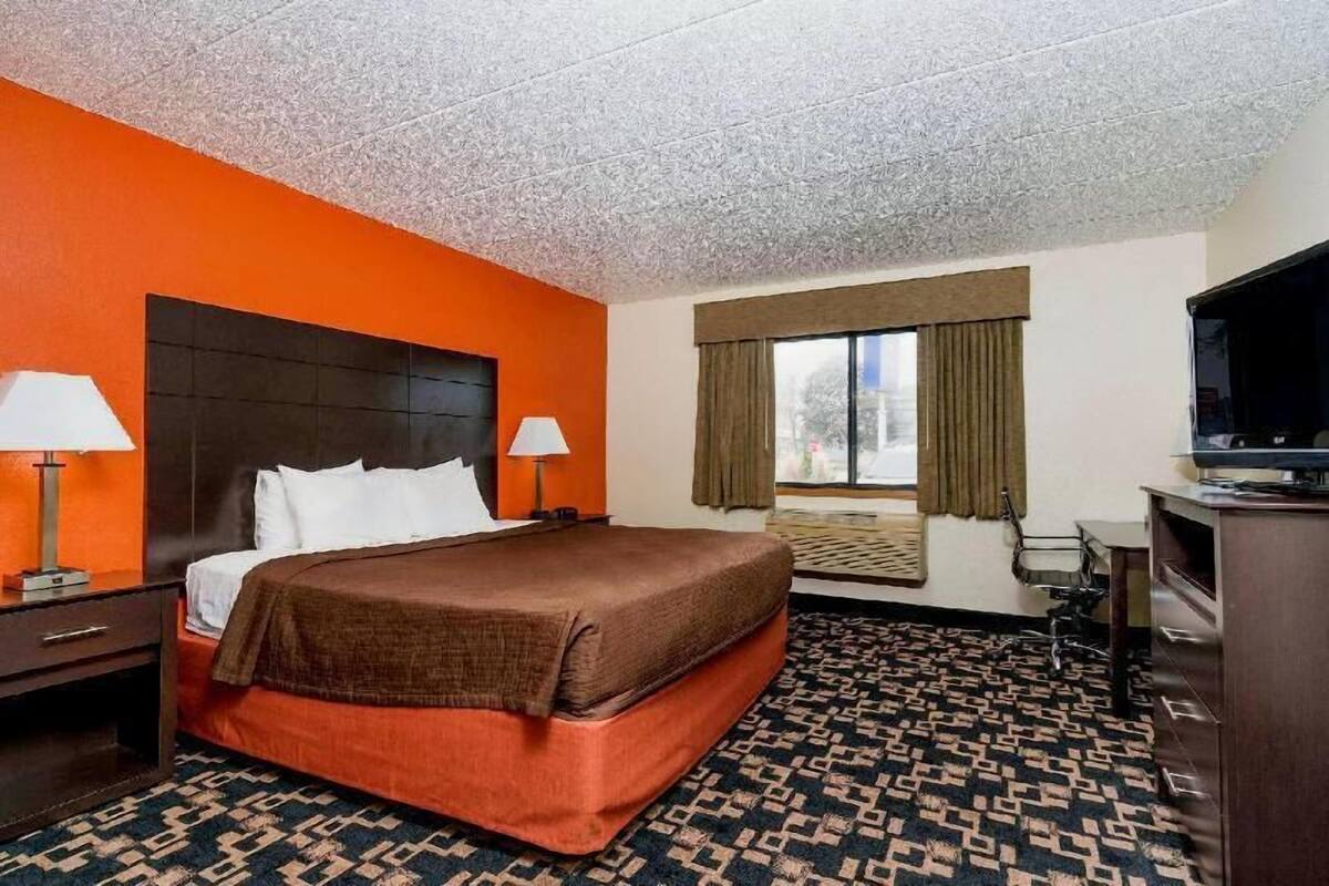 Ein Hotelzimmer mit einem großen Bett, zwei Nachttischen mit Lampen, einem Fernseher, einem Fenster mit Vorhängen und einem gemusterten Teppich.