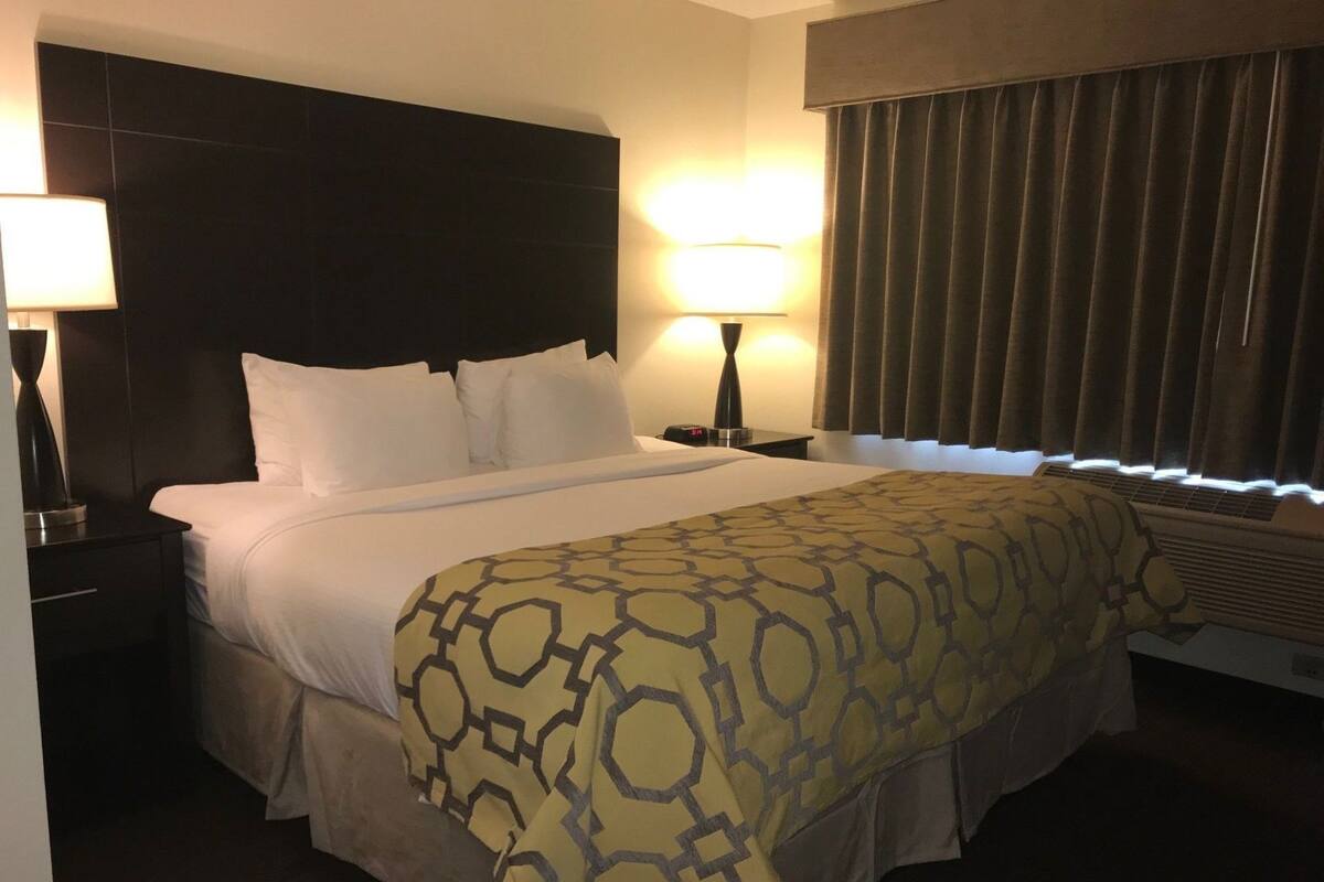 Ein Hotelzimmer mit Bett, Nachttisch, Lampe, Fenster mit Vorhängen und Klimaanlage.