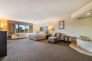 Quarto de hotel com cama, escrivaninha, cadeira, mesinha, sofá, luminária, quadro na parede e janela com cortinas.