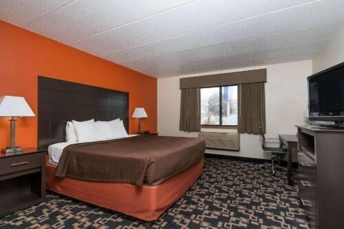 Ein Hotelzimmer mit einem Bett, zwei Nachttischen mit Lampen, einem Schreibtisch mit Stuhl, einem Fernseher und einem Fenster mit Vorhängen.