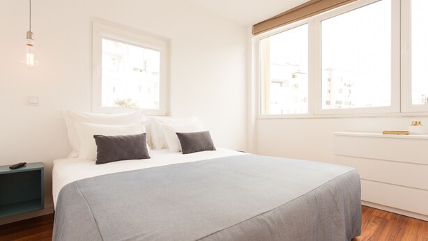 Une chambre moderne avec un grand lit, une table de chevet, une fenêtre donnant sur des immeubles et une petite table de nuit avec une lampe.