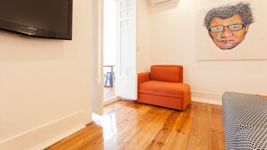 Une pièce avec un téléviseur à écran plat fixé au mur, un tableau au mur, un fauteuil orange et un canapé à motifs.