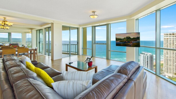 A spacious living room with a large sofa, a coffee table, and a TV mounted on the wall. The room has large windows offering a view of the ocean and a cityscape.