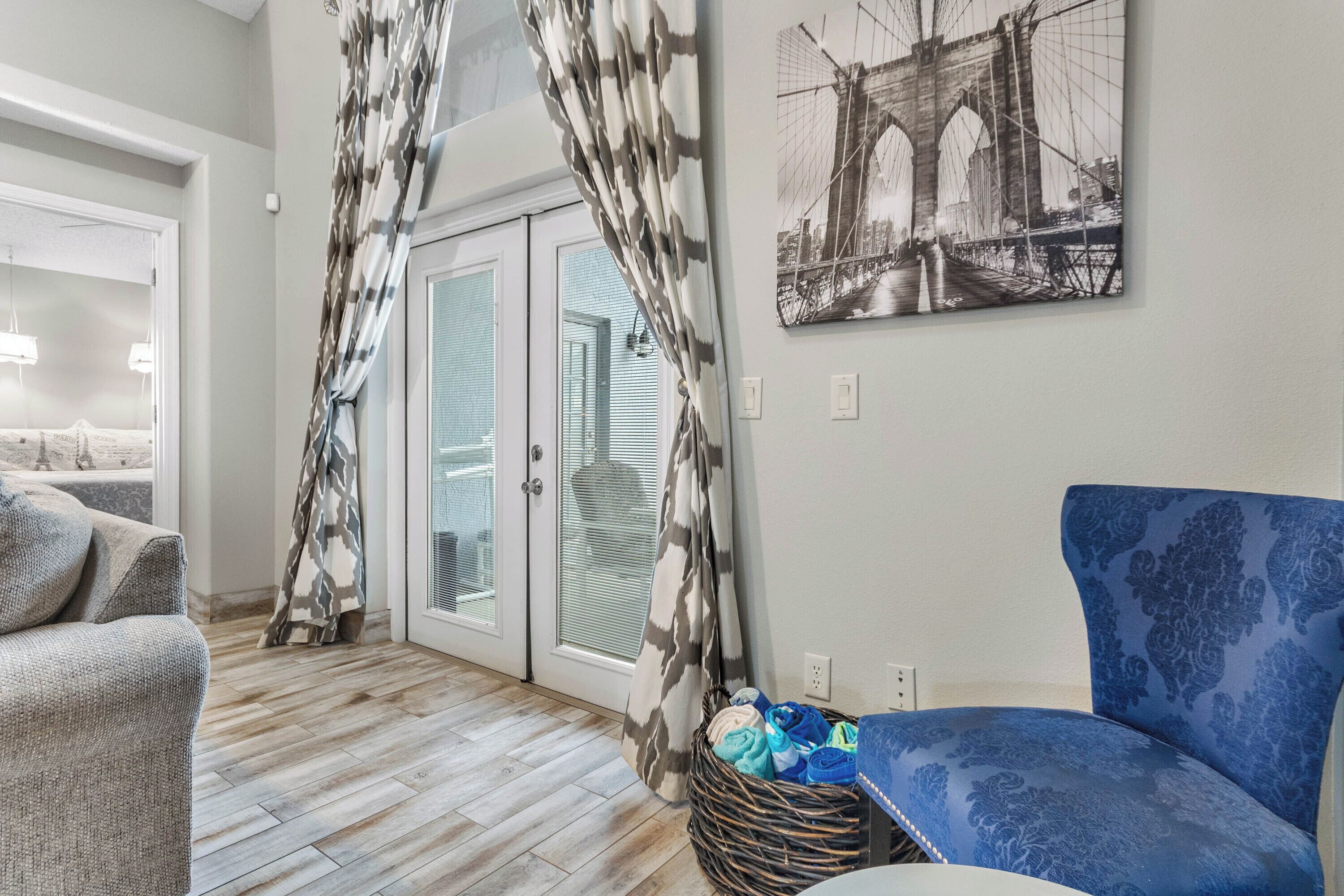 Un salon avec un canapé bleu à motifs, un canapé gris, un panier en osier contenant des vêtements pliés et une porte ornée de rideaux à motifs.