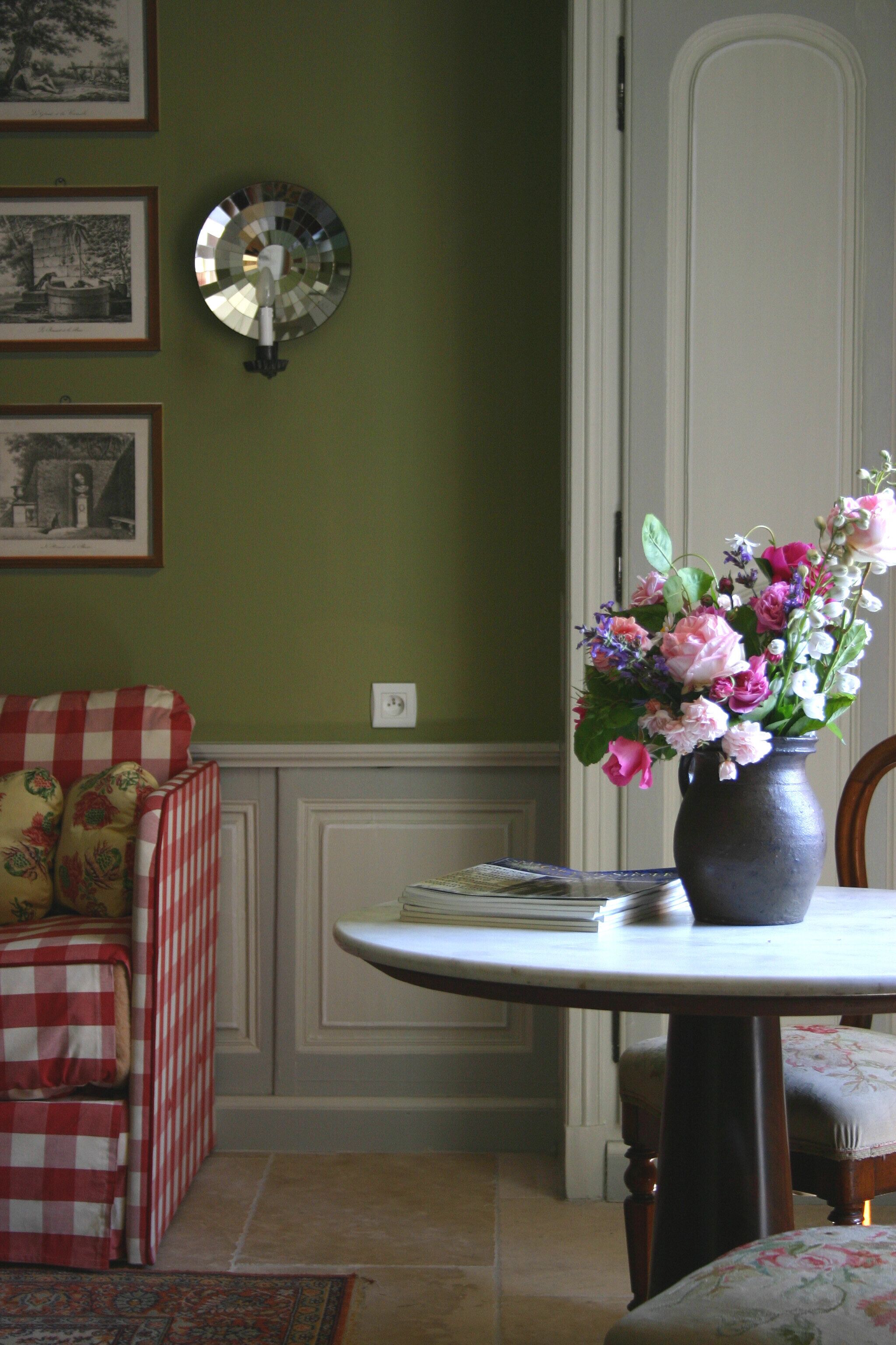 Una habitación con una pared verde, un sofá a cuadros, una mesa redonda con un florero y cuadros enmarcados en la pared.